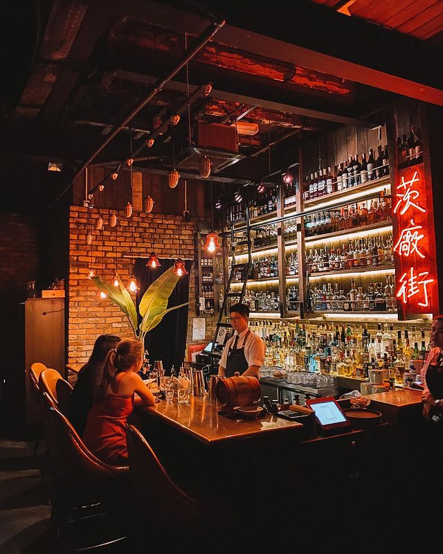 a cocktail bar with a bartender serving two seated guests with cocktails